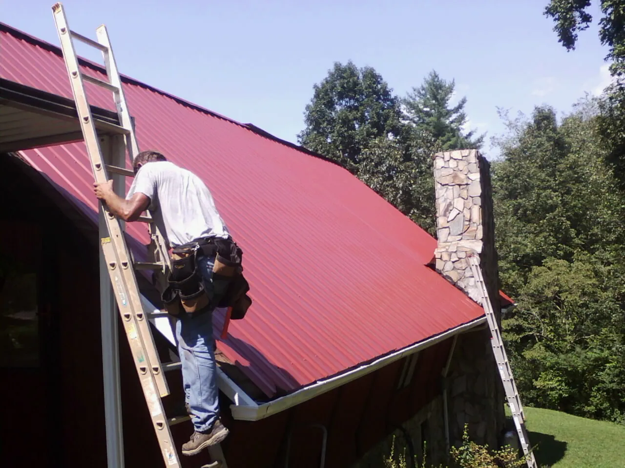 Roofer on a ladder beside a red metal roof and stone chimney — Soffit & Fascia Repair by Roofing Brothers in Forked River